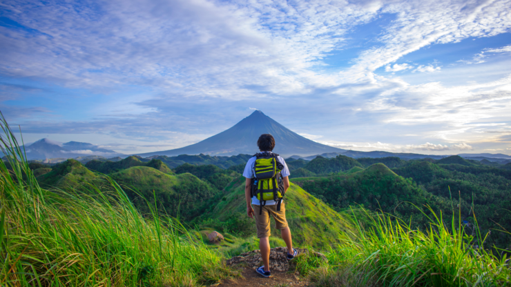 Man Wearing White Shirt, Brown Shorts, and Green Backpack Standing on Hill