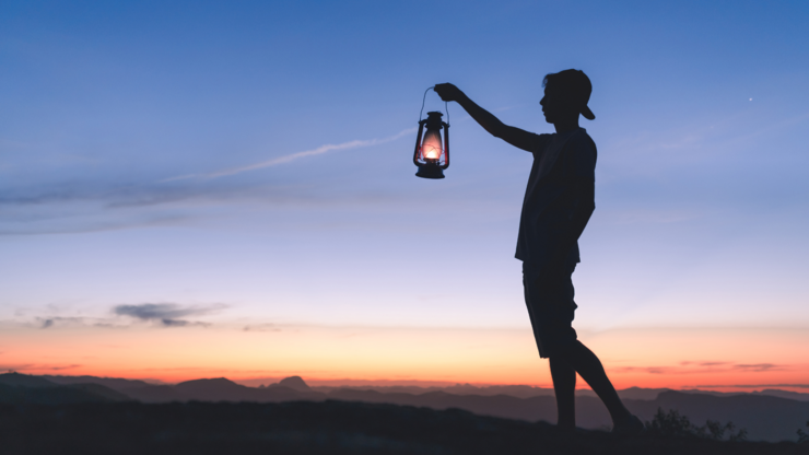 Silhouette of a man holding a lit kerosene lamp to light his way in the dark, with sky after sunset in the background.