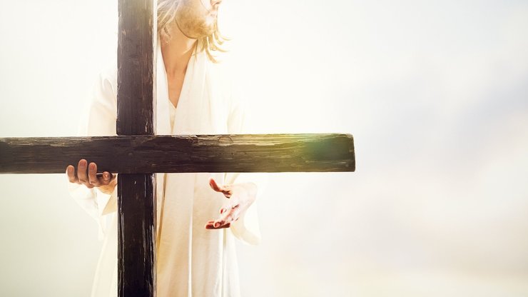 A representation of Jesus Christ in an idyllic heaven-like setting, extending his hand with the invitation of new life, his other hand holding the cross of crucifixion.  His face is obscured, and bright golden light shines around him. 